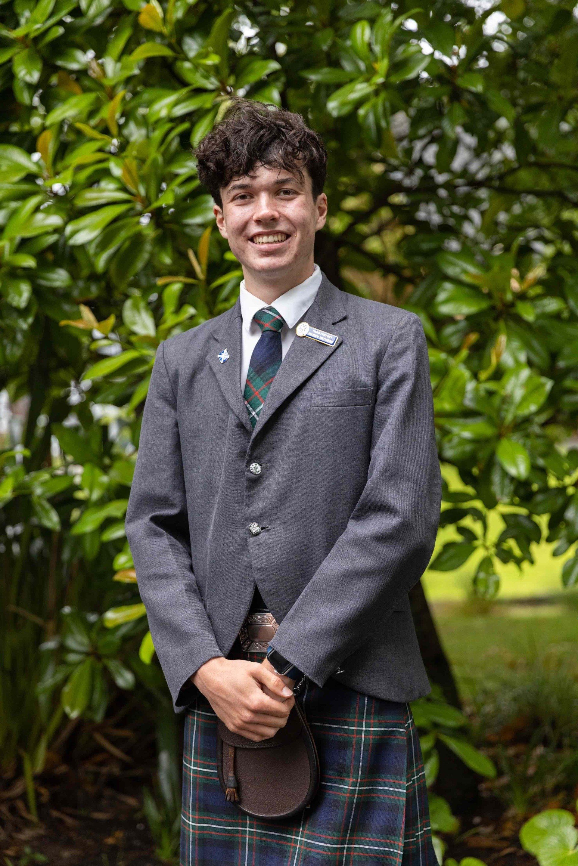 A young man in a suit and kilt is standing in front of a tree.