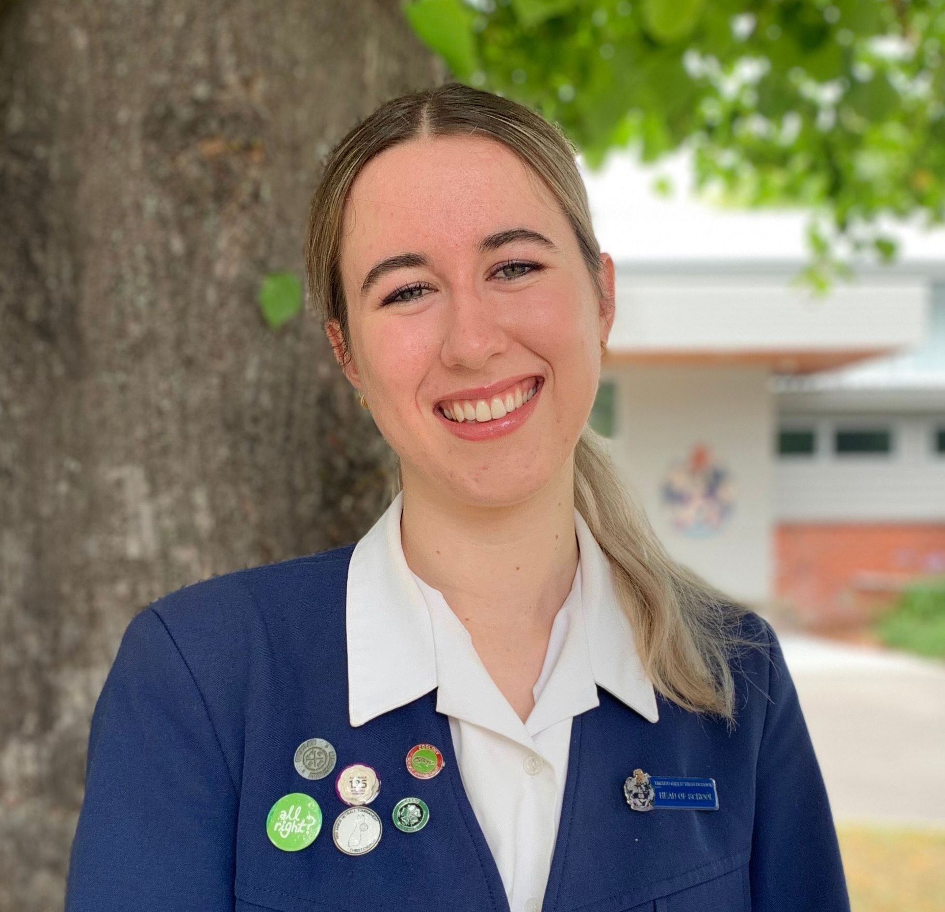 A woman in a blue jacket and white shirt is smiling in front of a tree.