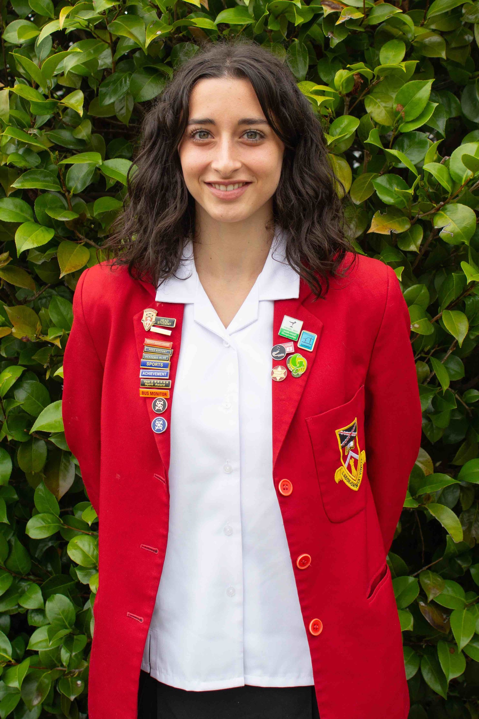 A woman in a red jacket and white shirt is standing in front of a bush.