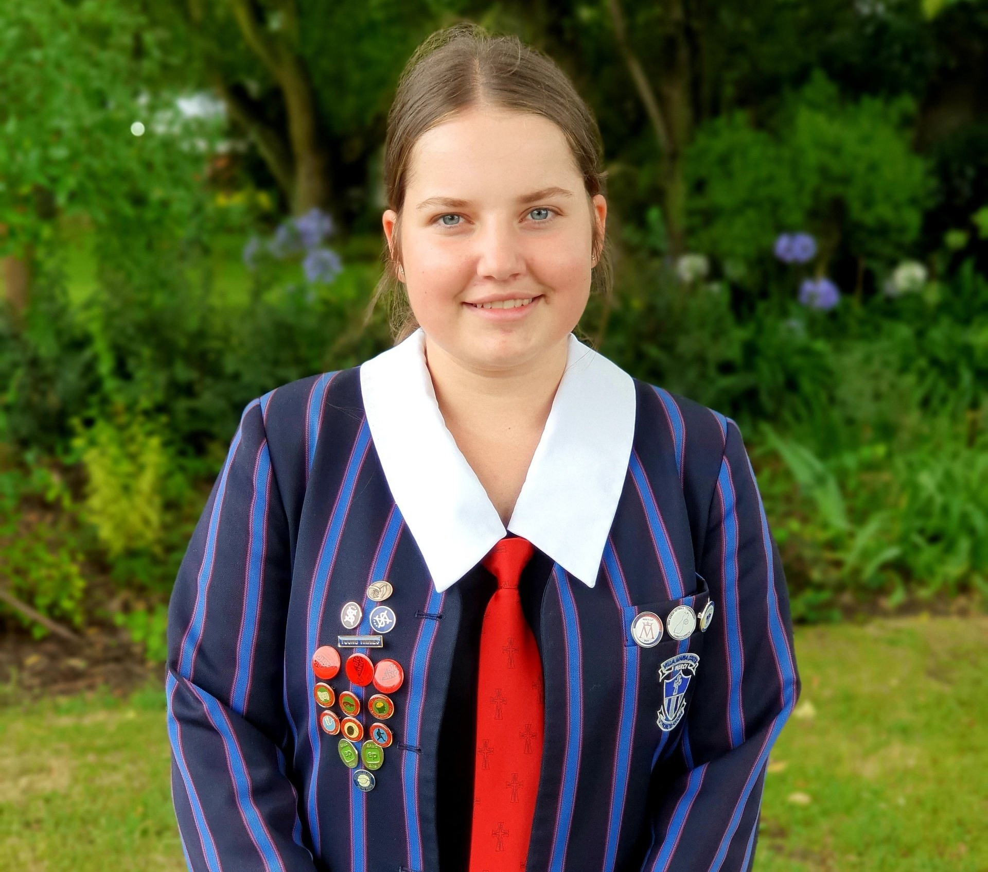 A young girl in a striped jacket and tie is standing in a garden.