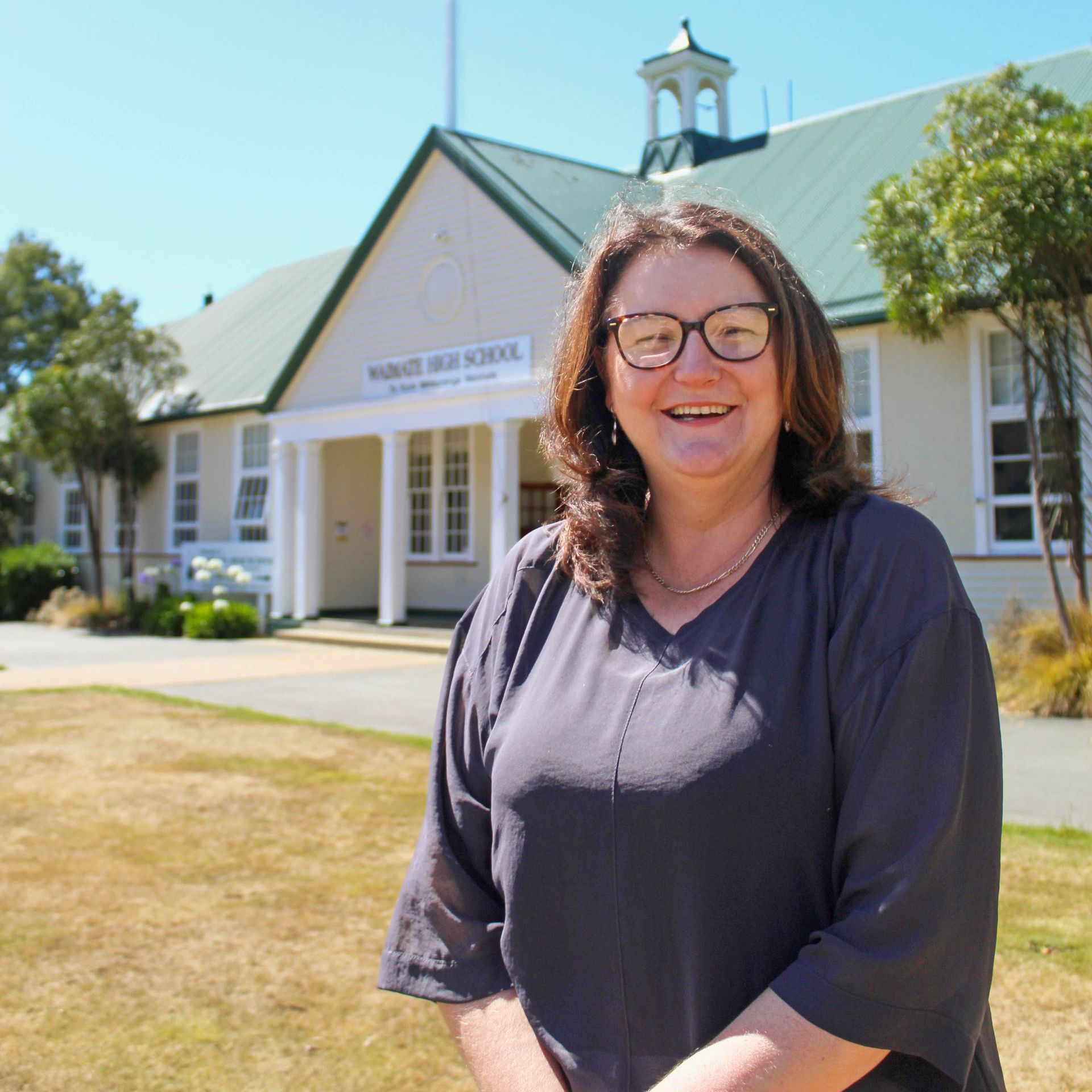 A woman wearing glasses stands in front of a school building