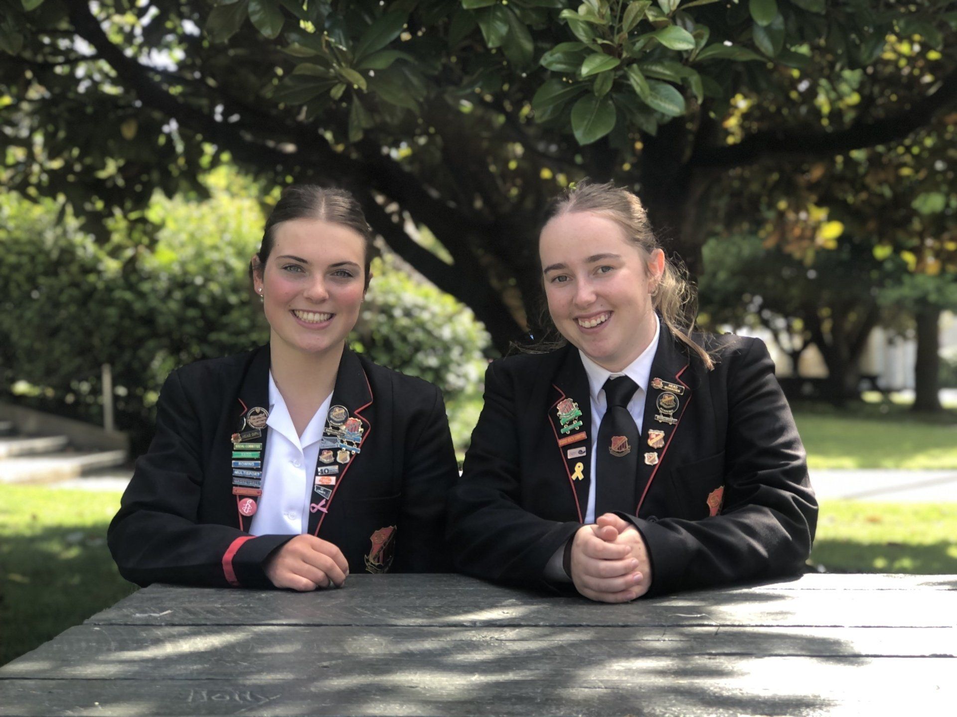 Two girls in school uniforms are sitting at a picnic table under a tree.