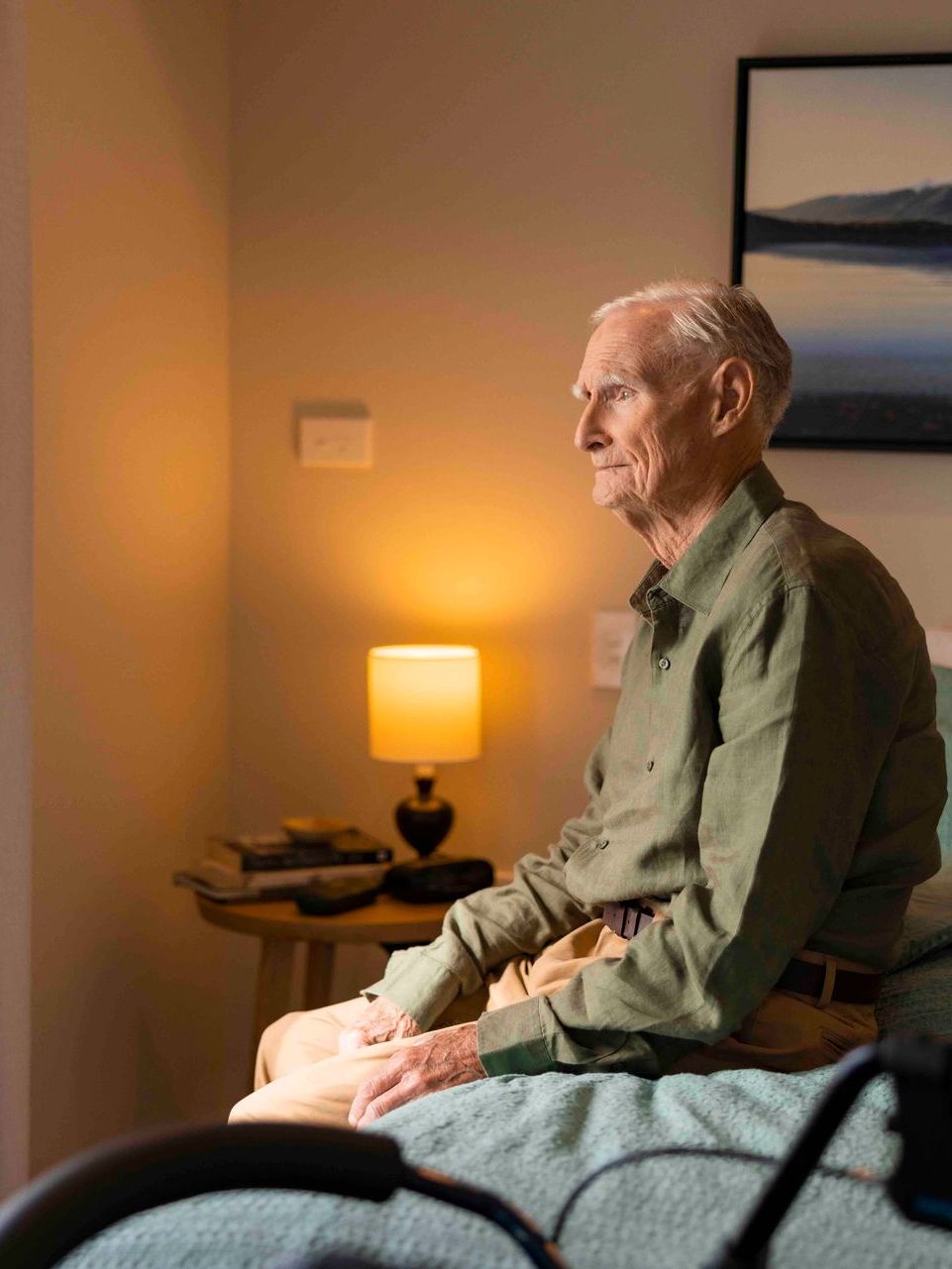 An elderly man is sitting on a bed looking out a window.