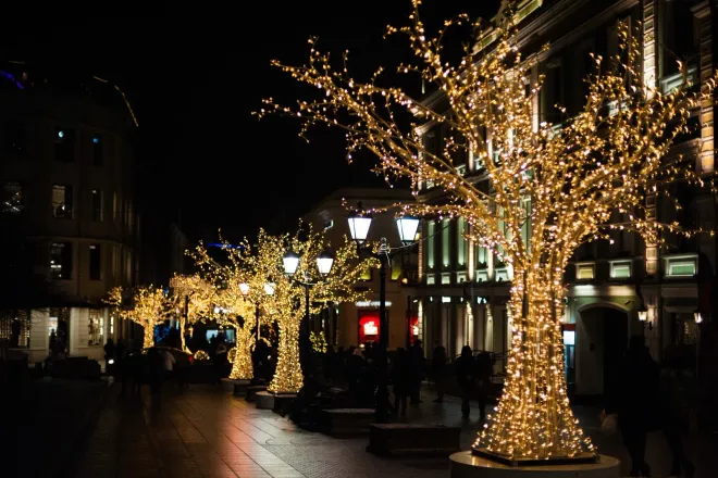 Trees wrapped in golden lights line a city street at night. Buildings and streetlights illuminate the scene.