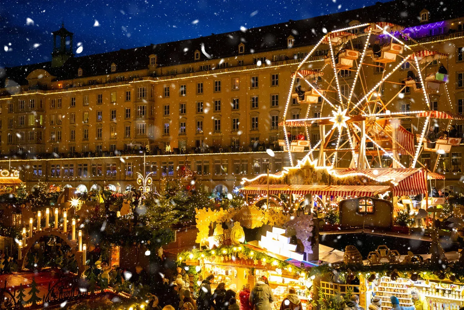 Christmas market with Ferris wheel, snow falling, buildings in the background at night.