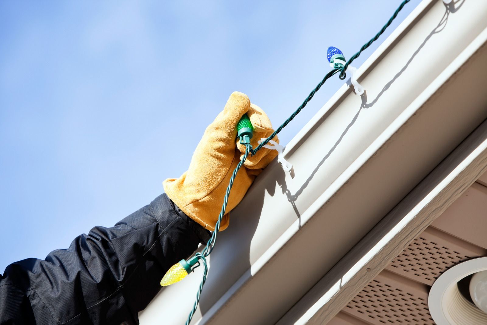 Woman on a ladder hanging Christmas lights on a house under a partly cloudy sky.