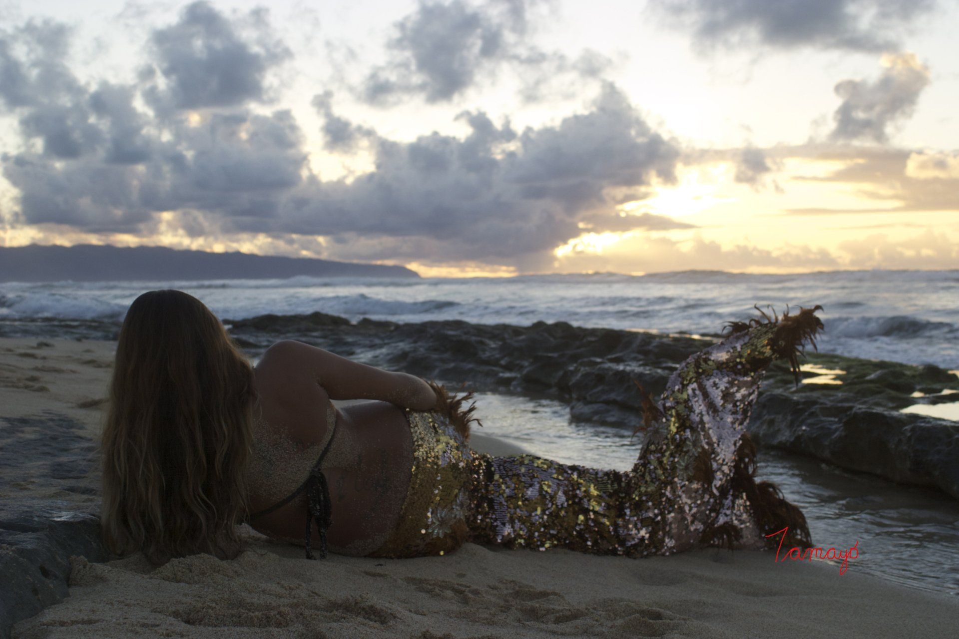 A woman in a mermaid costume is laying on the beach at sunset.