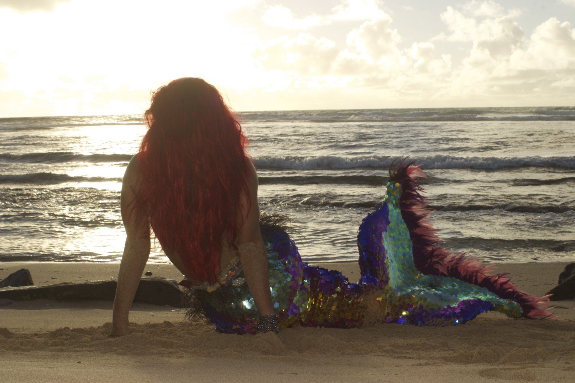 A woman in a mermaid costume is sitting on the beach looking at the ocean.