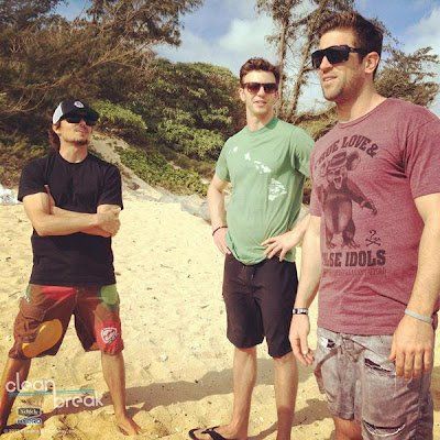 Three men standing on a beach with one wearing a shirt that says the idols