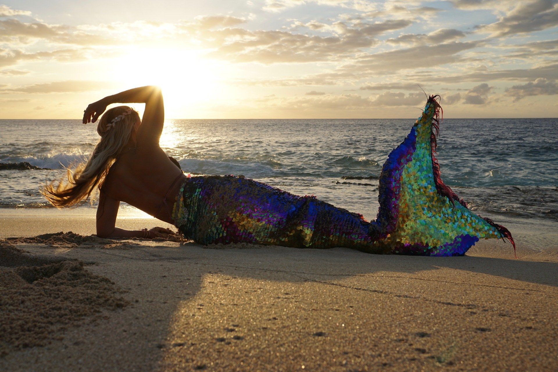 A mermaid is laying on the beach at sunset.