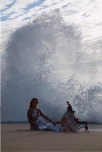 A woman is sitting on the beach in front of a wave.