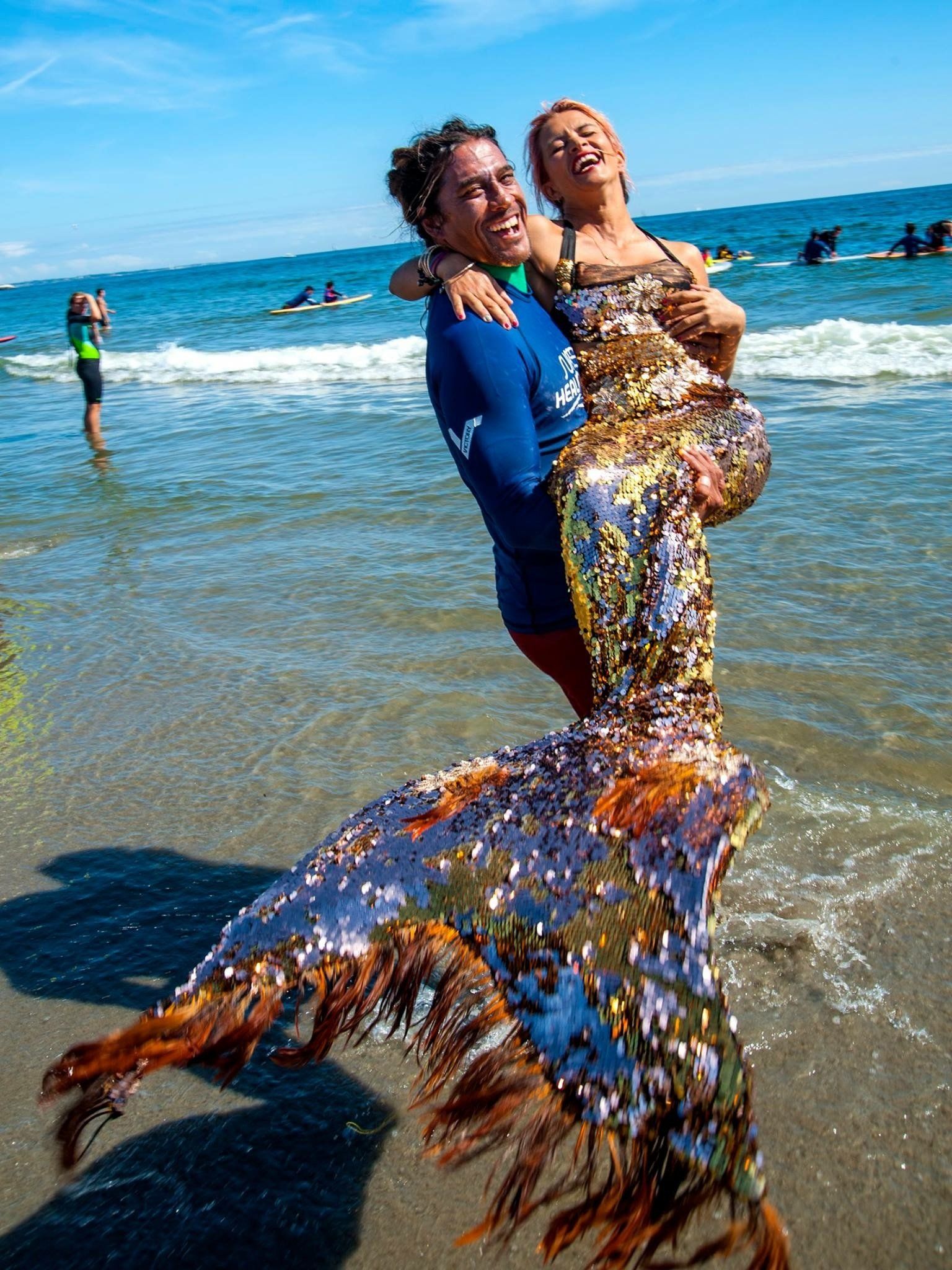 A man is holding a woman in a mermaid costume on the beach.