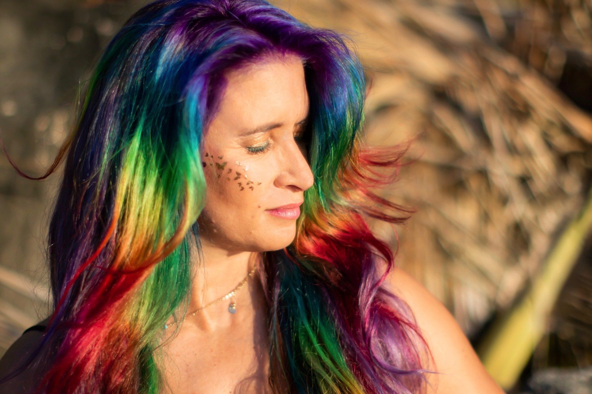 A woman with rainbow hair is standing on the beach with her eyes closed.