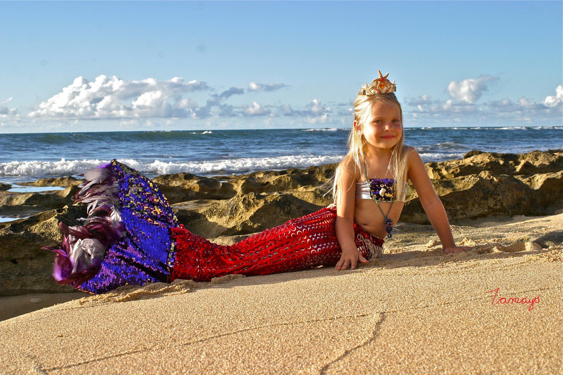 A little girl dressed as a mermaid is laying on the beach.