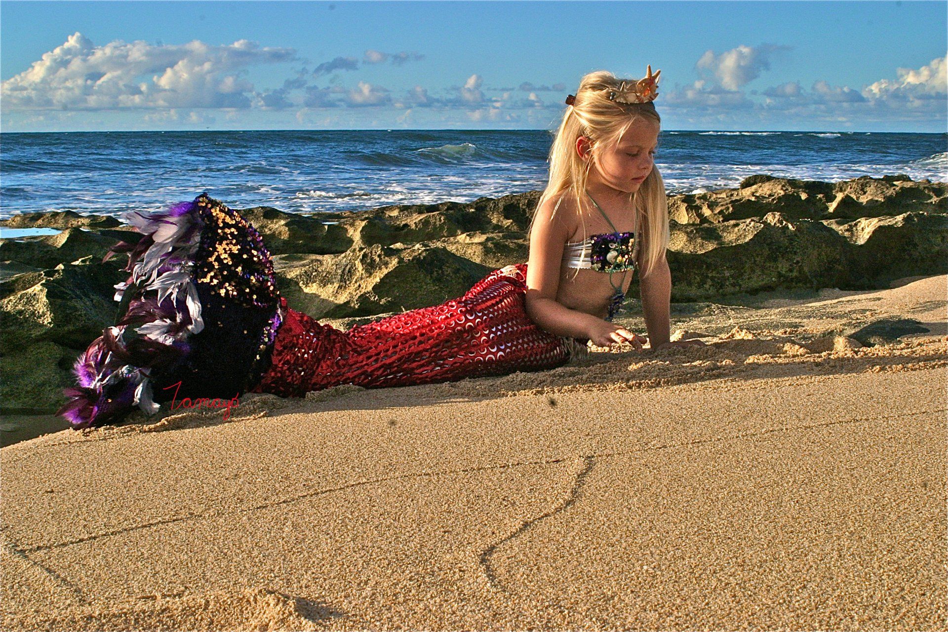 A little girl dressed as a mermaid is laying on the beach