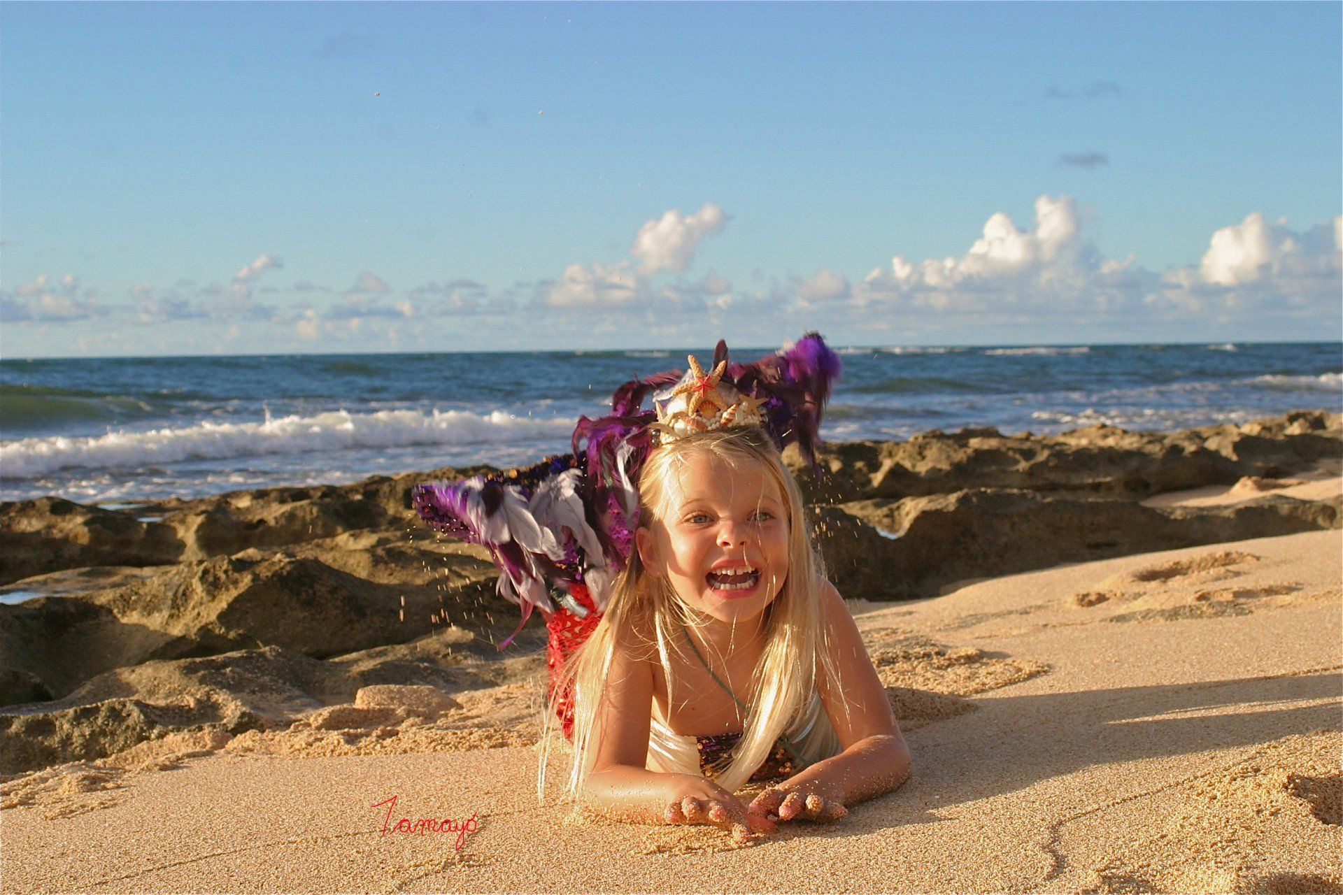 A little girl dressed as a mermaid is laying on the beach