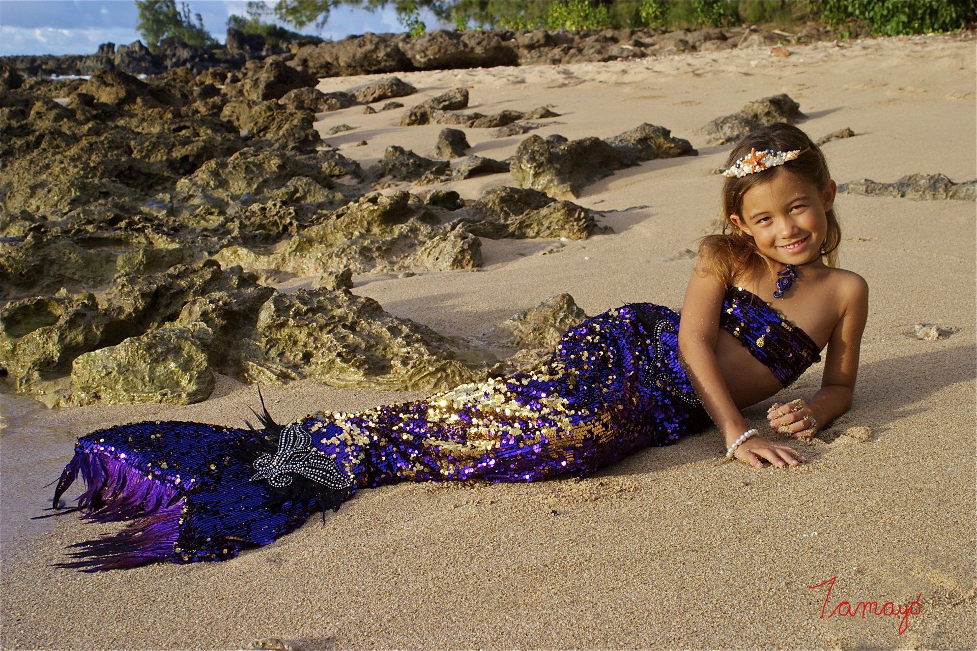 A little girl dressed as a mermaid is laying on the beach