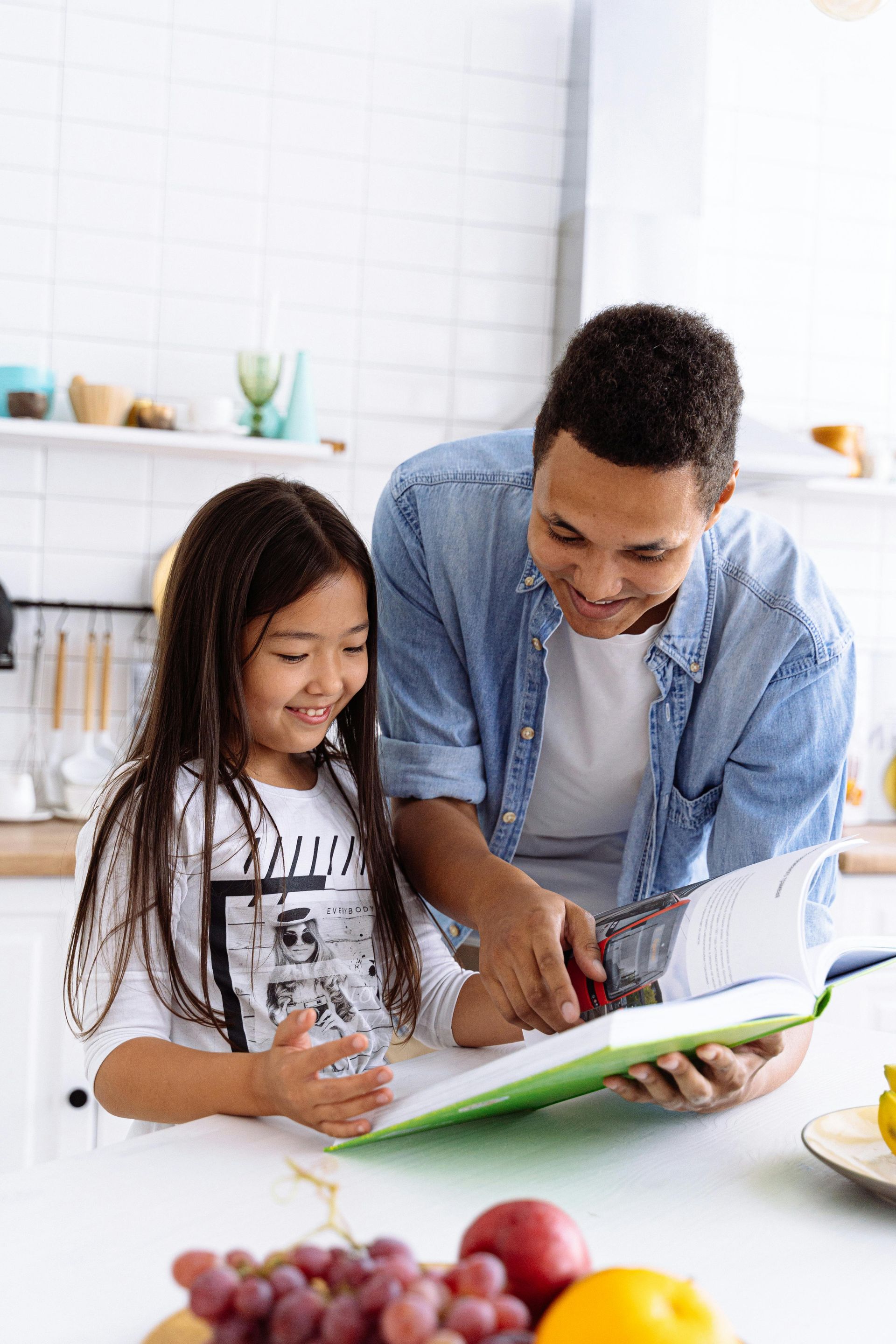 A man and a child looking at a cookbook in a kitchen, smiling. Fruit on the table.