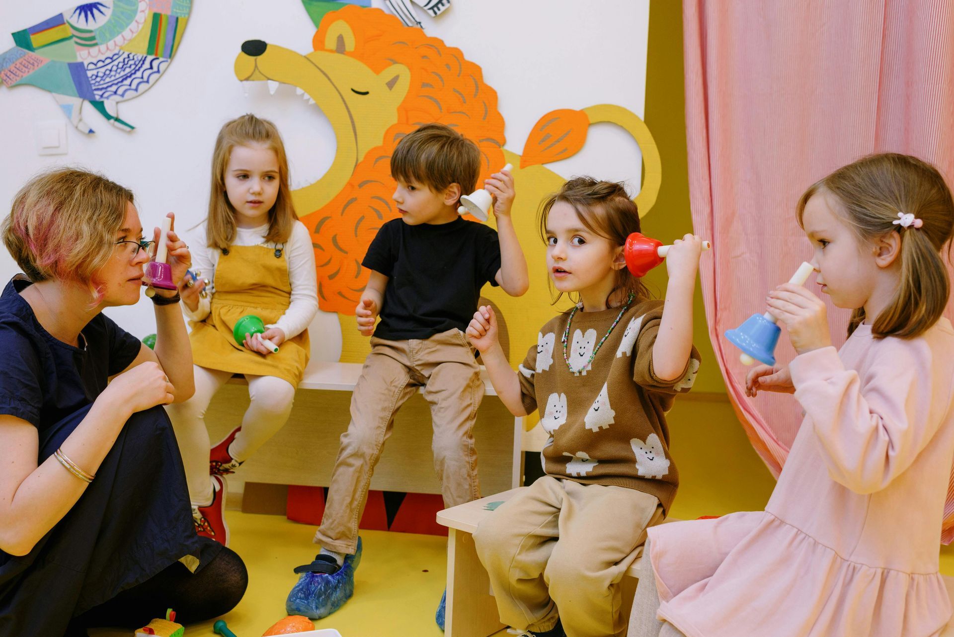 Children in a room playing with musical instruments, teacher assisting, colorful decorations.