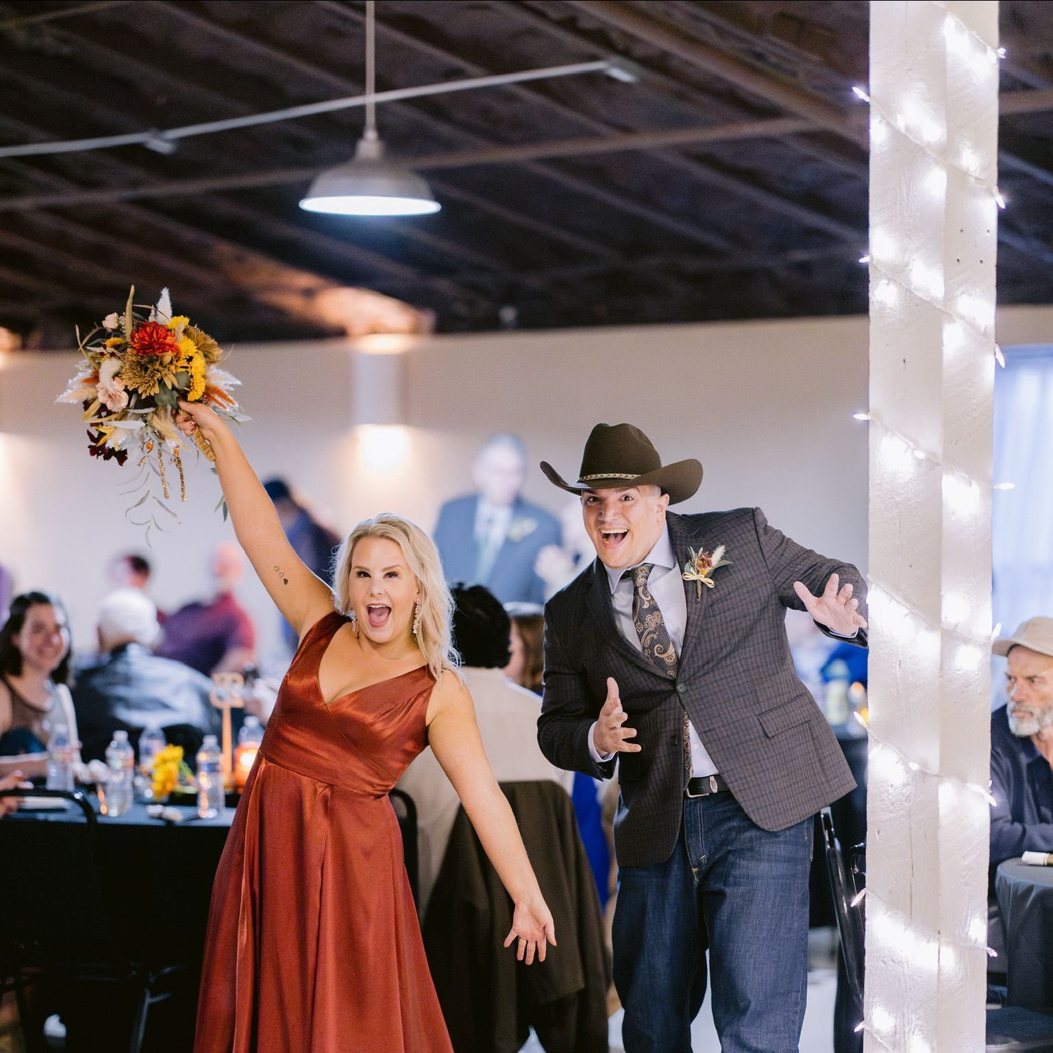 a woman in a red dress is holding a bouquet of flowers while standing next to a man in a cowboy hat at their wedding reception