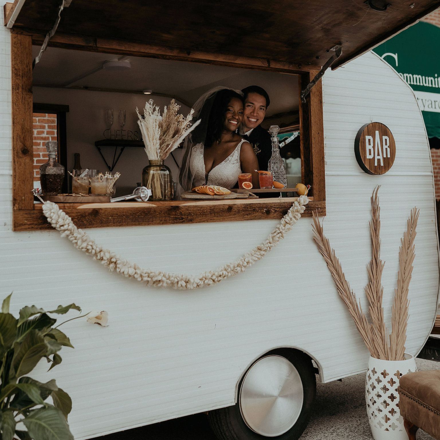 a bride and groom are posing in a food truck that is decorated in white and brown plants, and is serving alcohol at their wedding