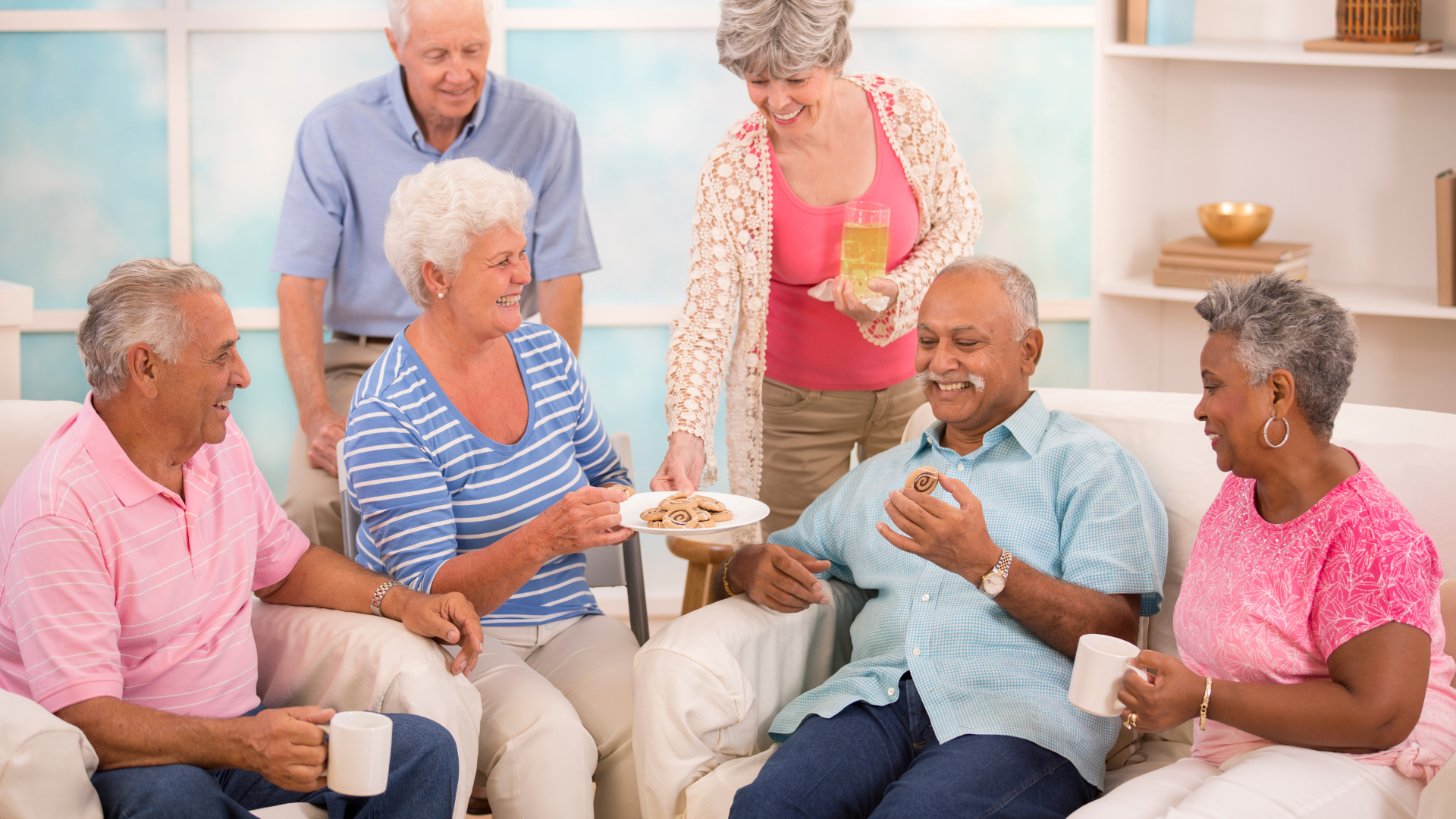 A group of senior adults socializing and sharing food and drinks on a sofa in a well-lit room.