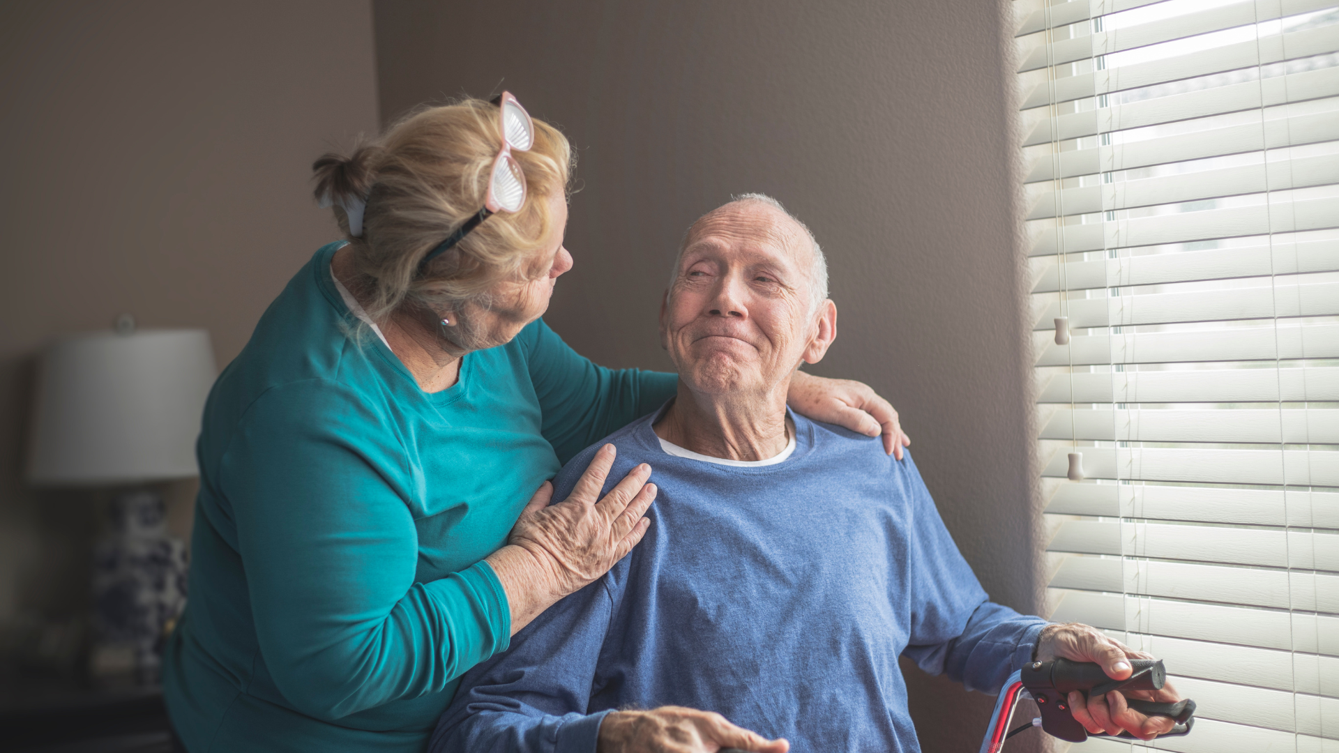 A woman in teal top puts a hand on the shoulder of an elderly man in blue, smiling at a window.