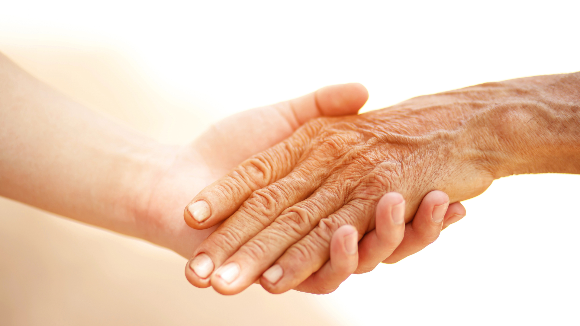 Two hands clasped together; one young, one old. The older hand is wrinkled and tan, clasped by a smooth, lighter-skinned hand.