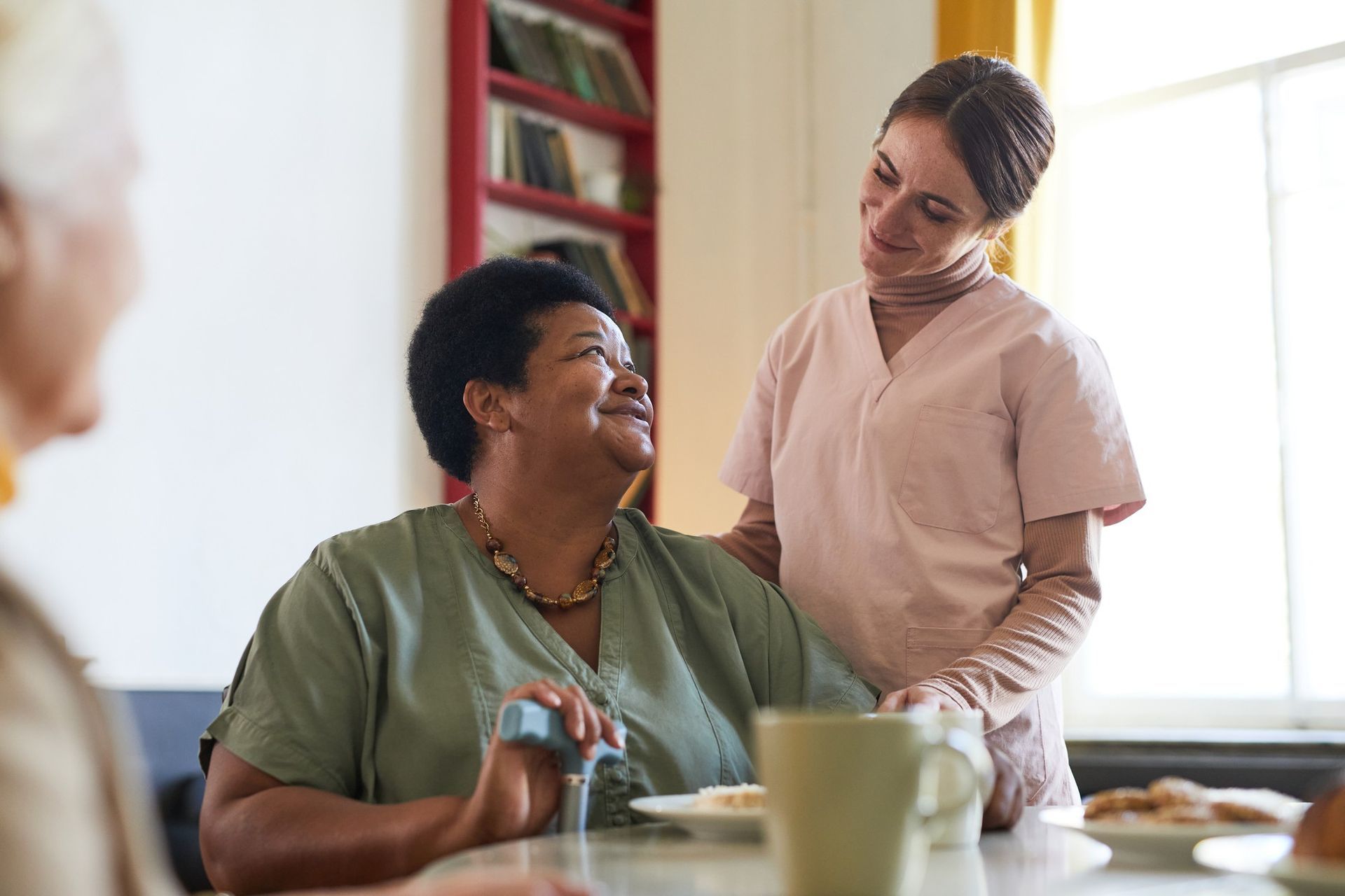 A caregiver comforts an elderly Black woman seated at a table, smiling and touching her shoulder; other person sits at the table.