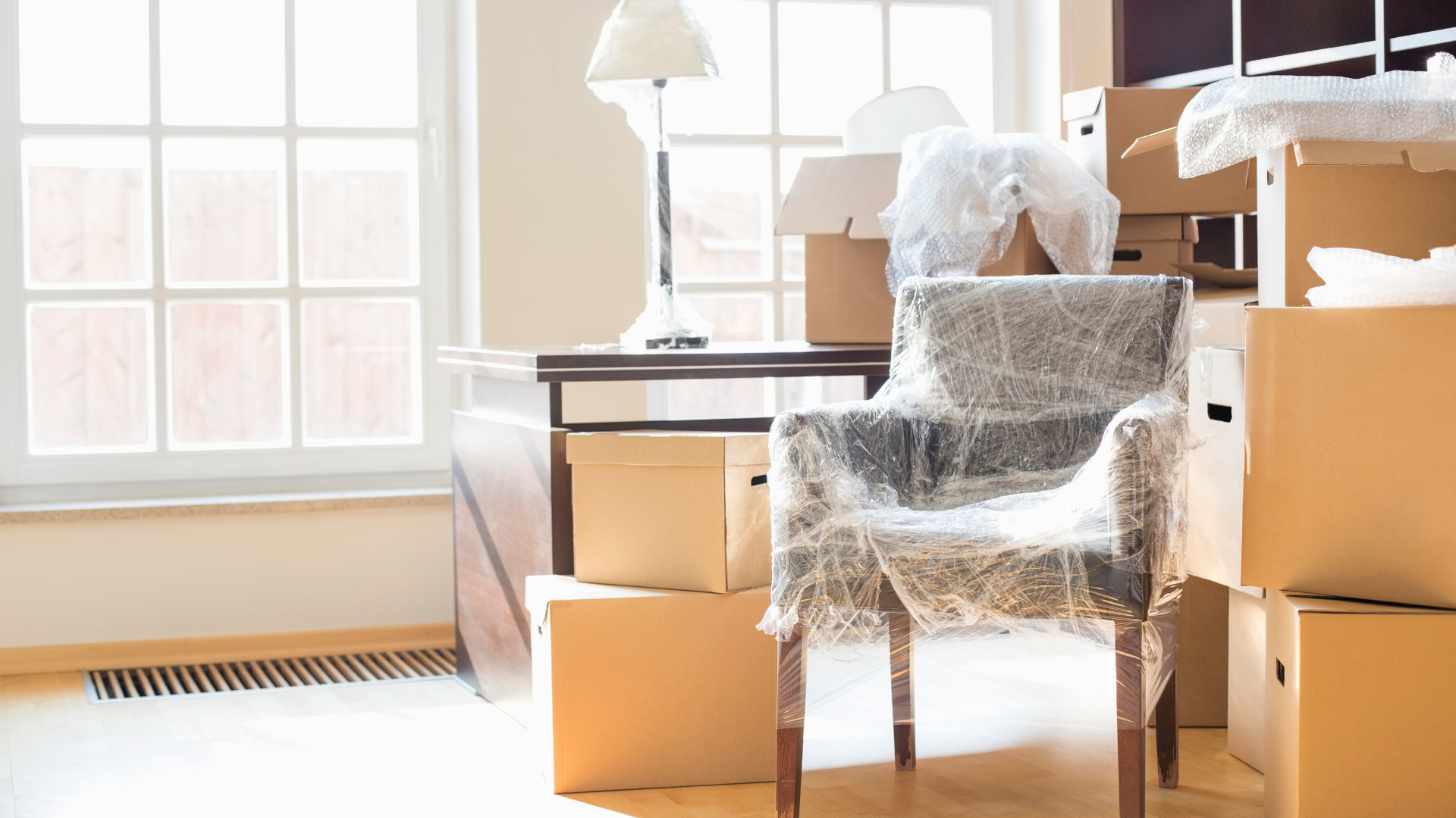 A room in disarray with moving boxes, a wrapped chair, and furniture by a bright window.