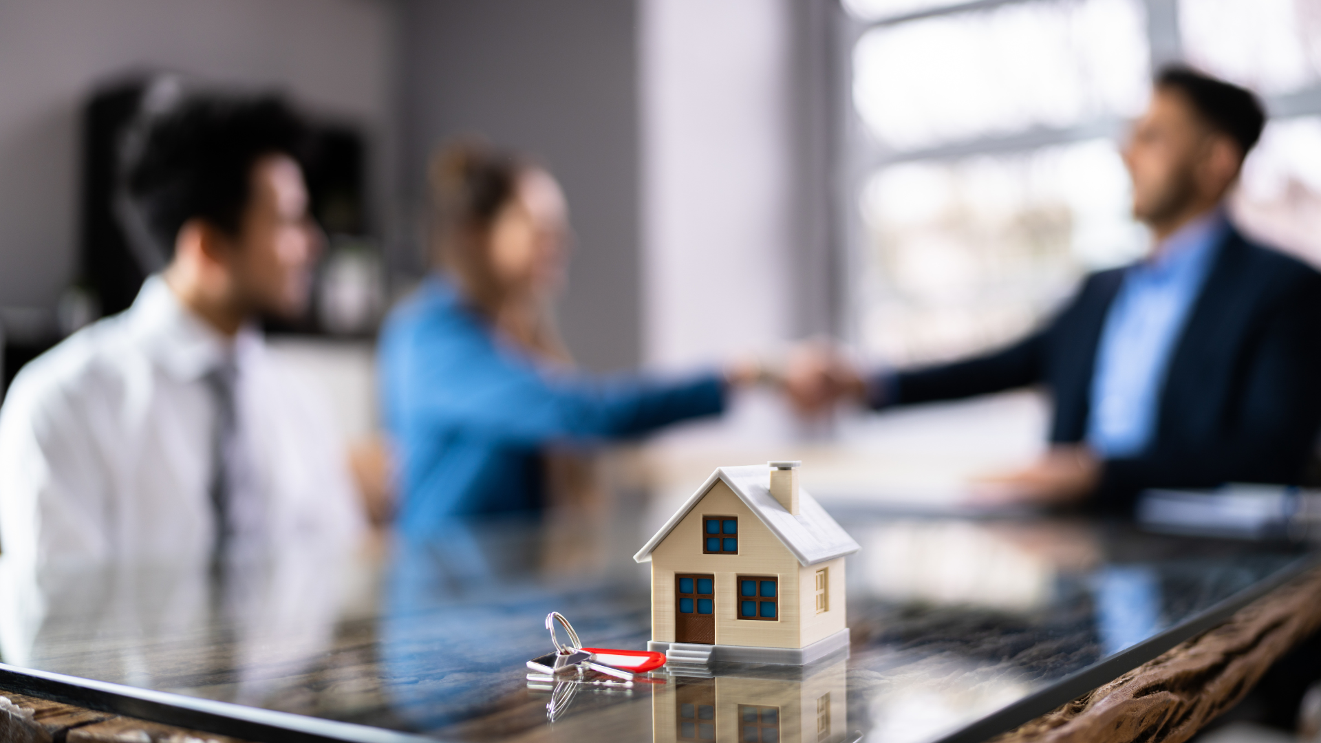 Miniature house with keys on a table; couple shaking hands with a realtor, blurred in the background.