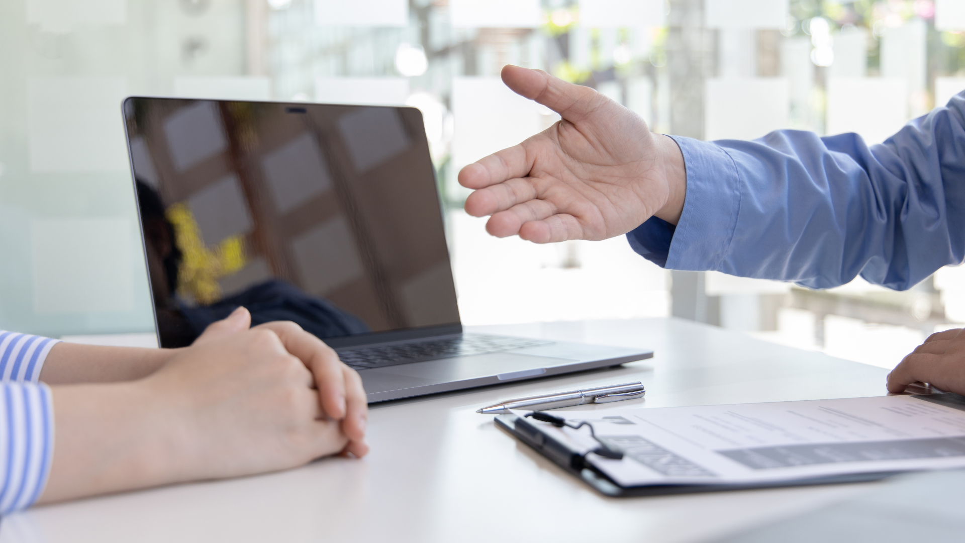 Two people at a desk; one extends a hand for a handshake, the other clasps their hands. Laptop and resume visible.