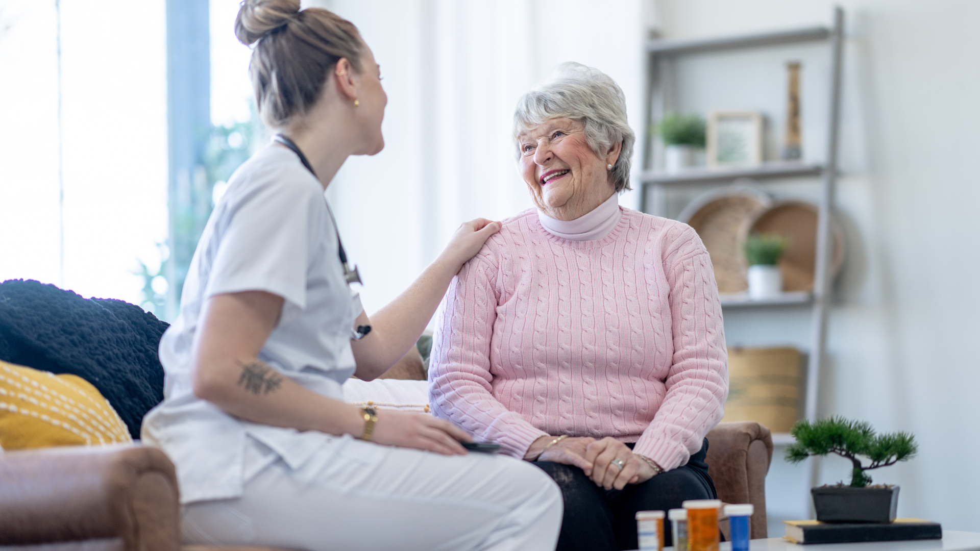 A healthcare worker smiles at an elderly woman sitting on a couch. The woman smiles back, with medicine bottles visible.