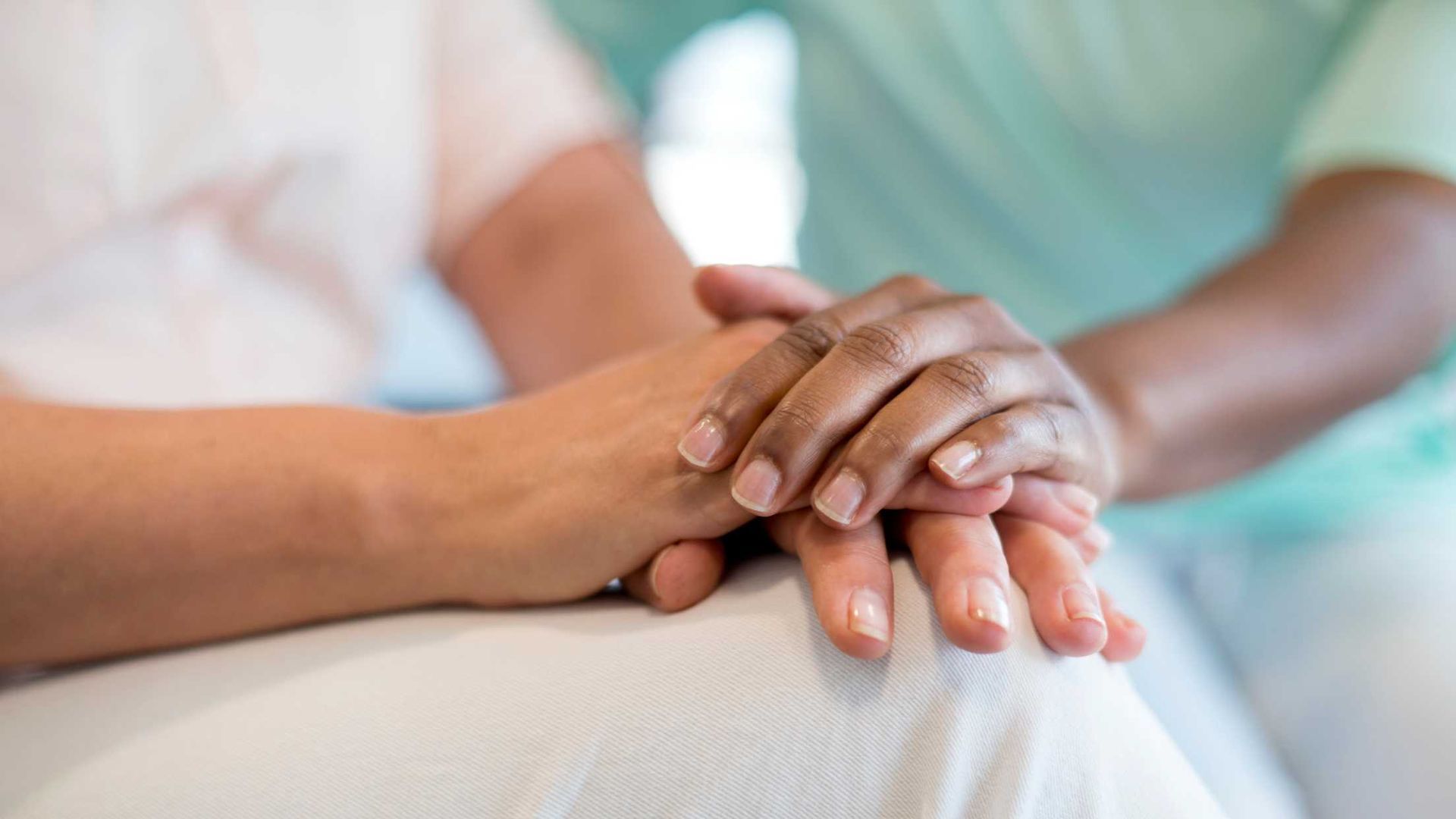 Hands of a light-skinned person are held by a darker-skinned person wearing a green top, offering comfort.