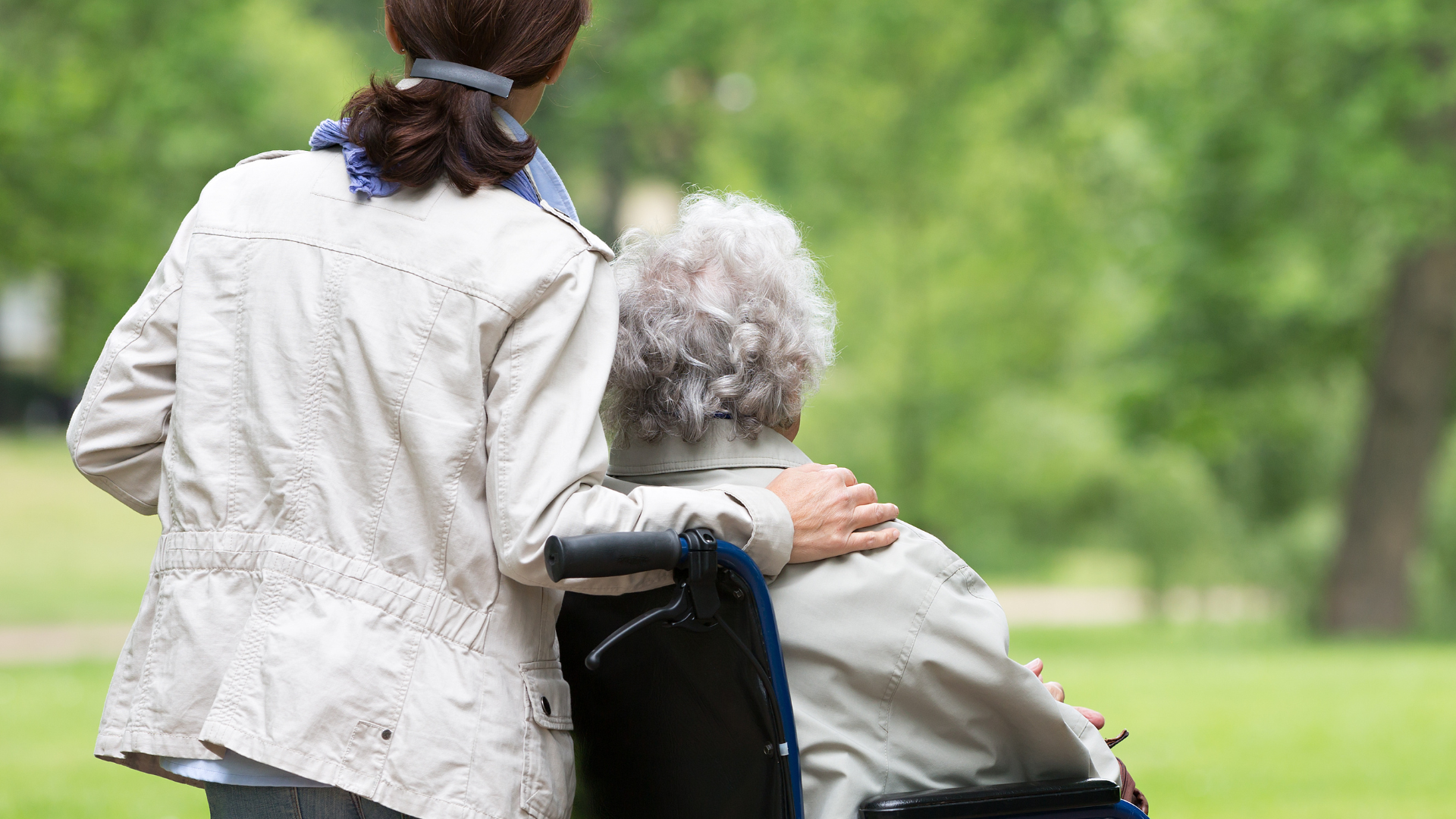 A younger woman in a cream jacket helps an elderly woman in a wheelchair outdoors.