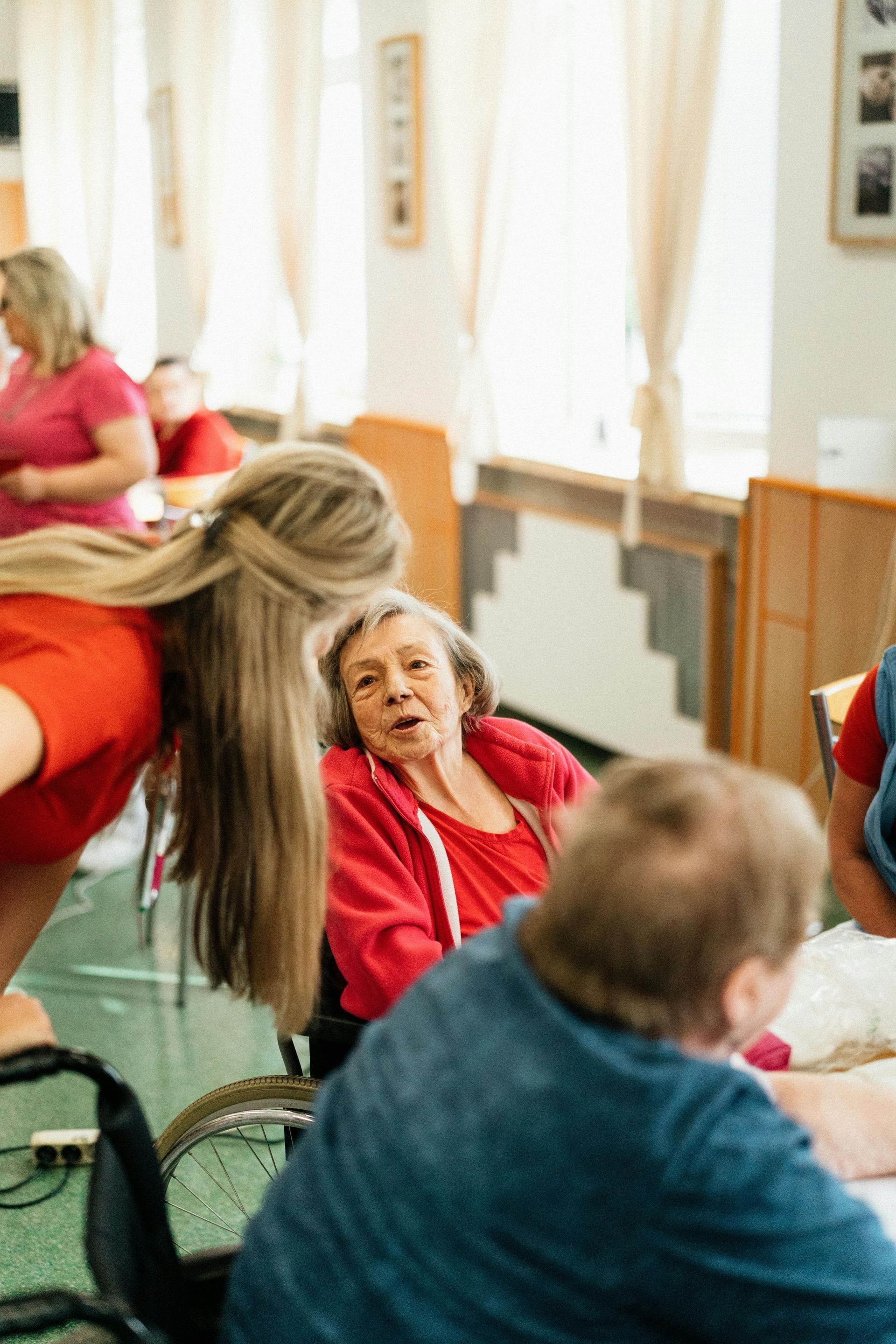 Woman in wheelchair smiles at a woman in red, as others socialize in a bright room with curtains and simple decor.