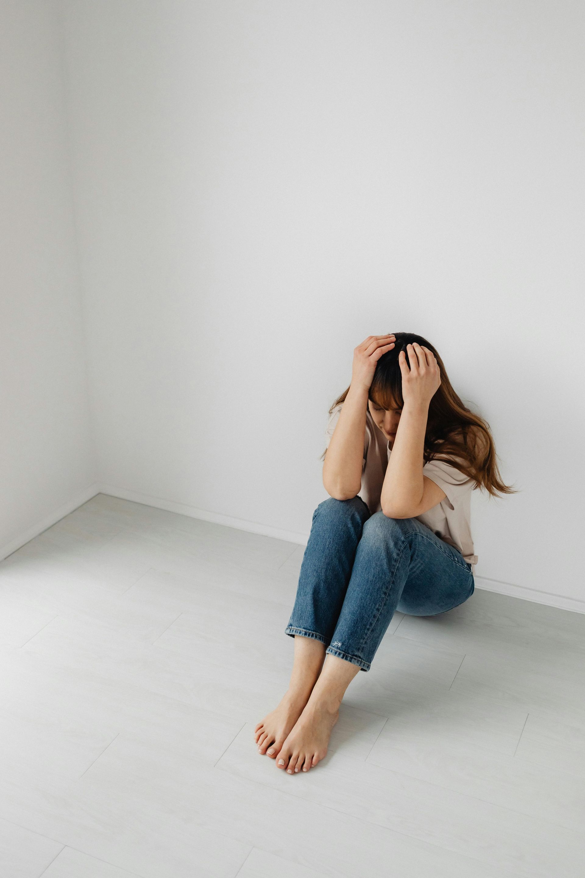 Person sits on floor, hands covering face, against a white wall in a corner; jeans, light-colored shirt.