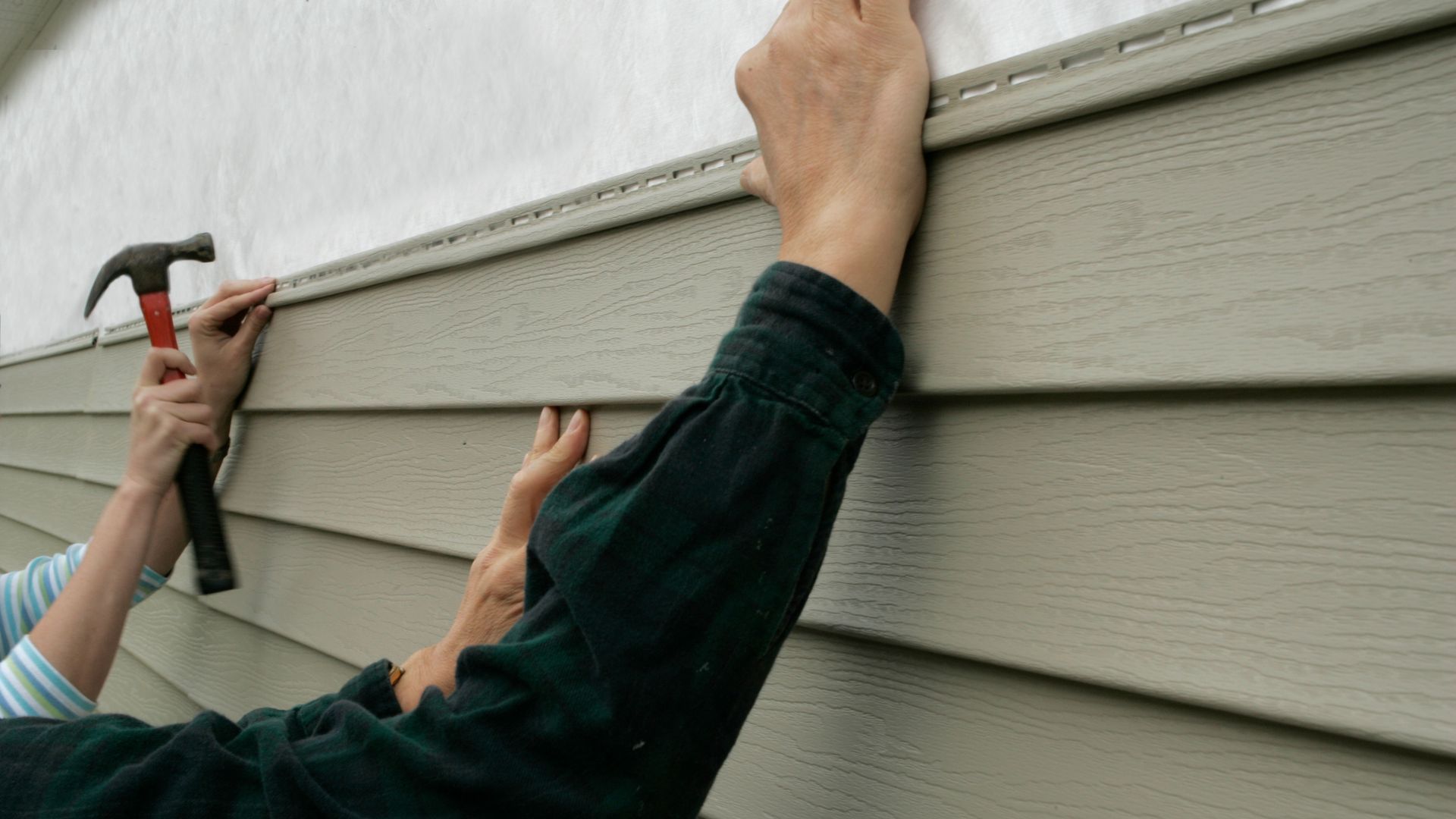 Hands hammering siding onto a house; light brown panels, blue shirt sleeves.