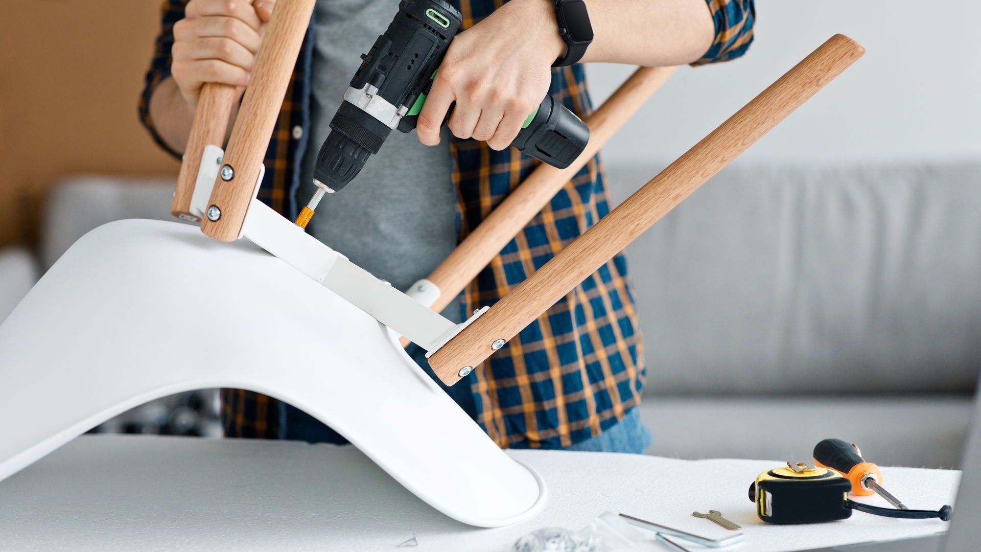 Person assembling a white chair with wooden legs using a power drill.