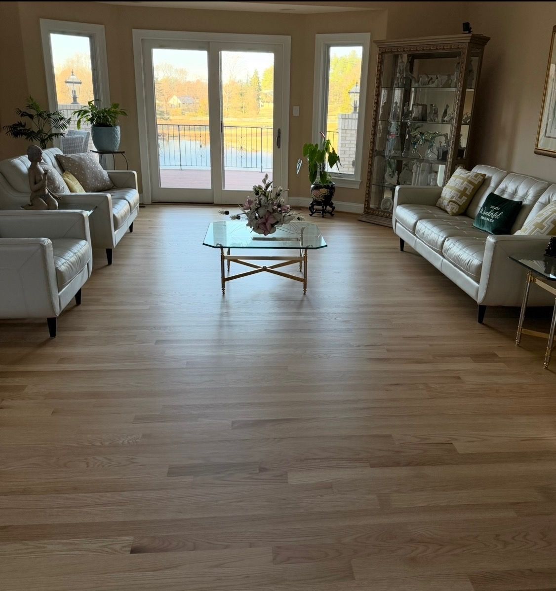 Living room with light hardwood floors, two white sofas, glass coffee table, and large glass doors.