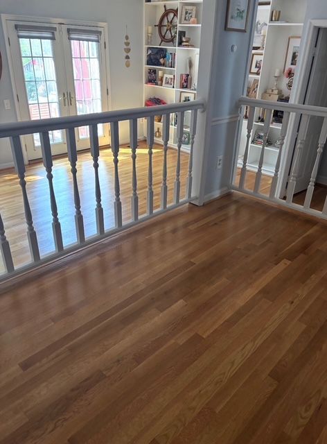 Wooden floors and white railing on a second-story landing overlook a sunlit room with built-in bookshelves.