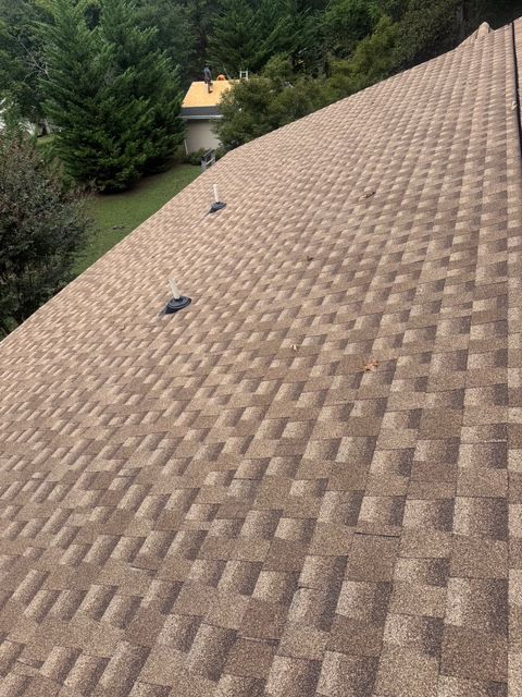 Close-up view of a brown asphalt shingle roof with three vents and surrounding trees.