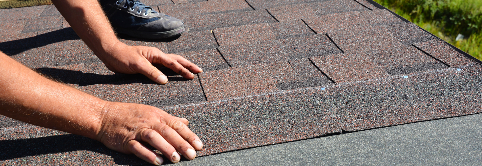 Hands installing asphalt shingles on a roof, visible setting: grass.