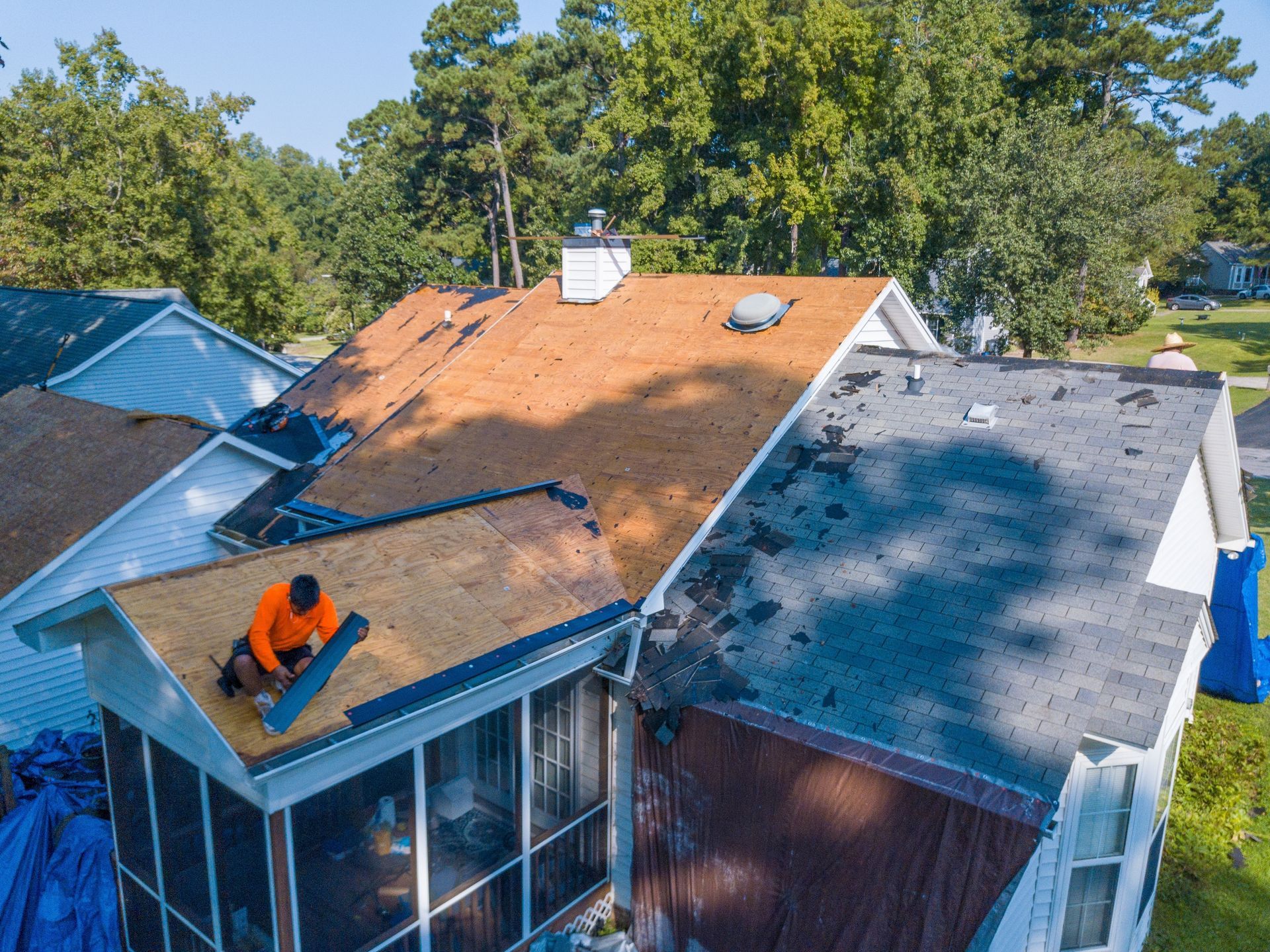 Roofer in orange shirt working on a roof partially covered with shingles. Houses and trees are in the background.