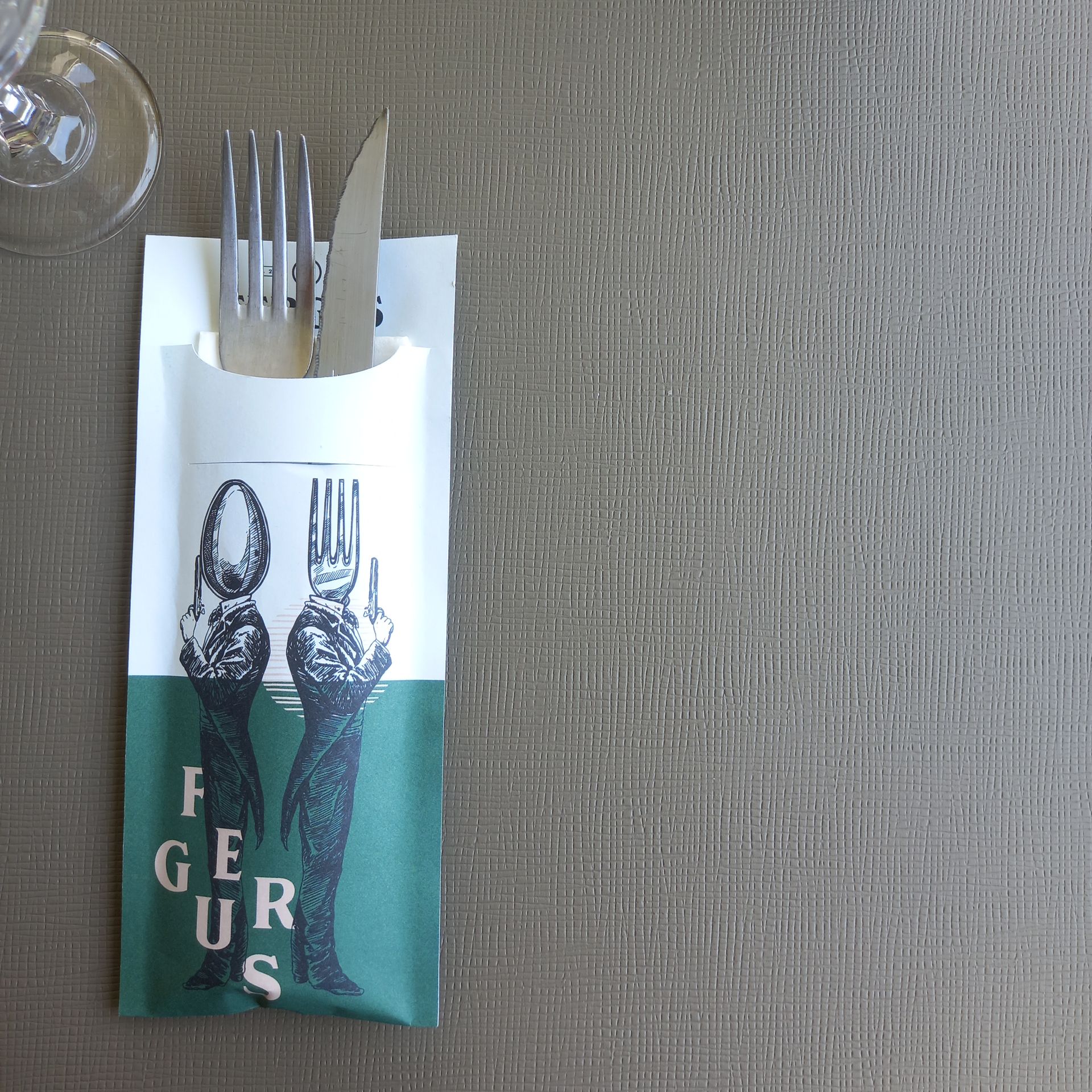 Une table dressée dans un restaurant avec un menu noir, des couverts, des verres et des serviettes roses.