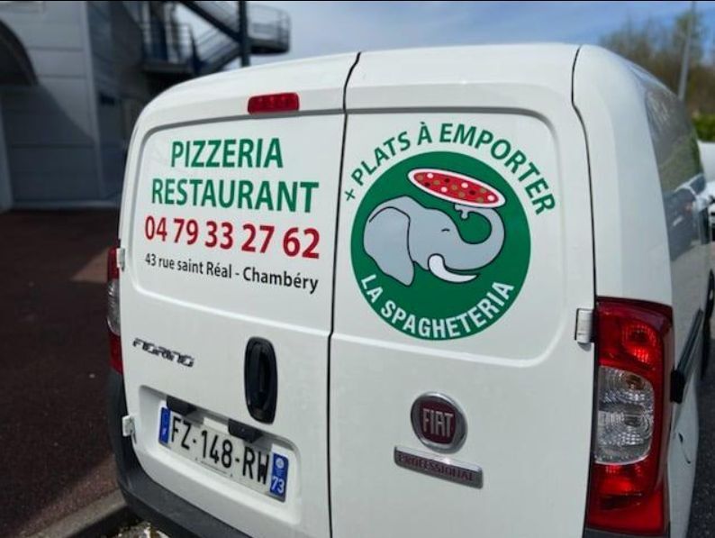Camionnette blanche avec logo de pizzeria : un éléphant tenant une pizza. Texte vert : « PIZZERIA RESTAURANT ». Numéro de téléphone et adresse. « Plats à emporter ».