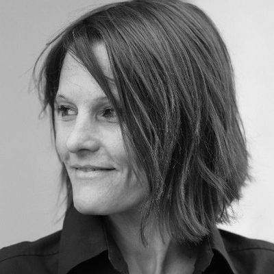 Femme aux cheveux courts sourit, regardant vers la gauche, en chemise noire, portrait en noir et blanc.