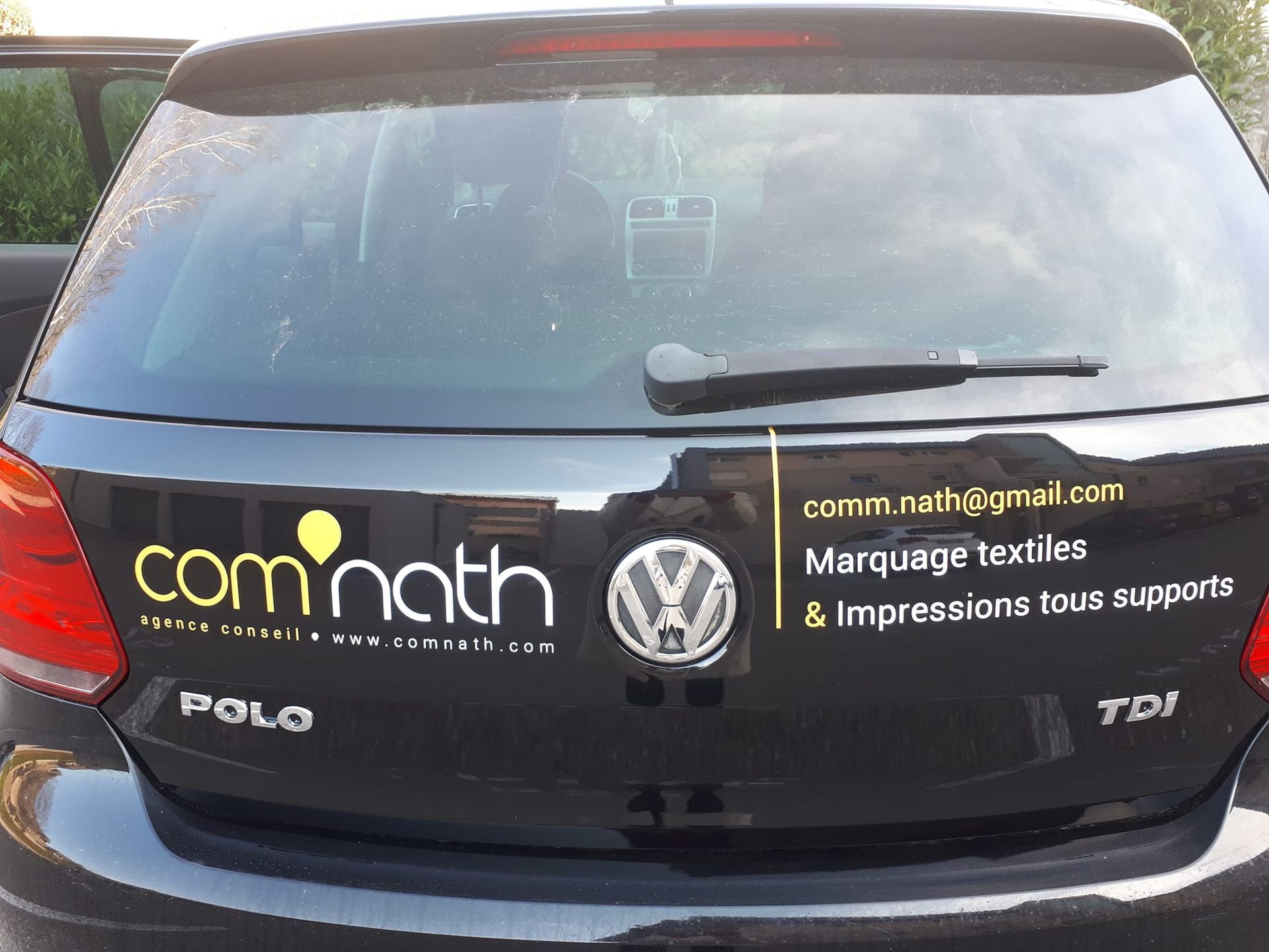 Voiture VW Polo noire avec logo de l'entreprise : « com.nath » et coordonnées sur la lunette arrière.