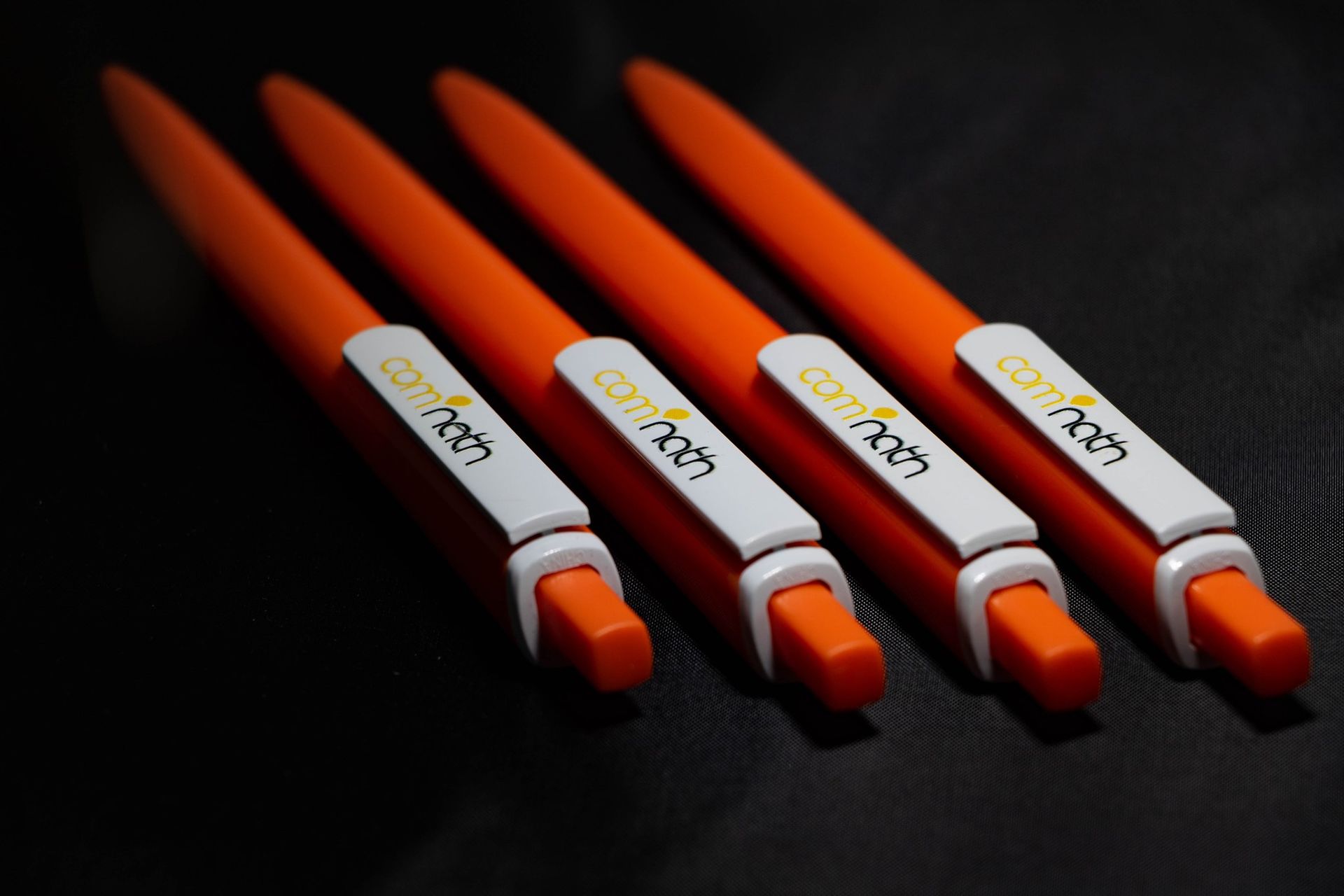 Quatre stylos orange avec clips rectangulaires blancs ; logo « comfinch » visible sur les clips.
