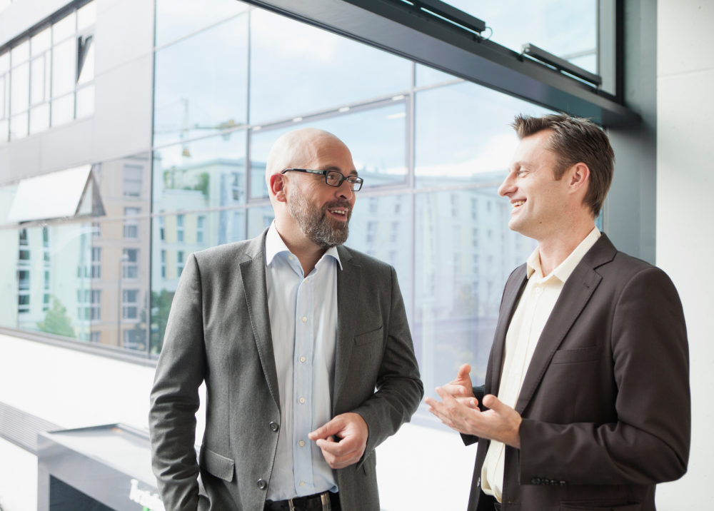 Two men in suits are talking to each other in front of a window.